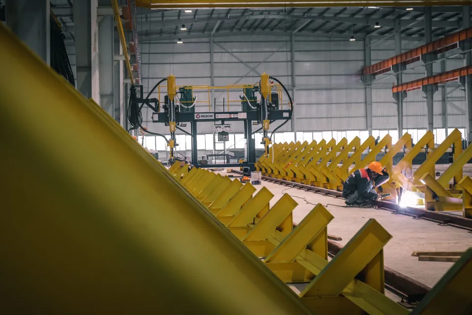 A worker welding metal in an industrial warehouse with heavy machinery.