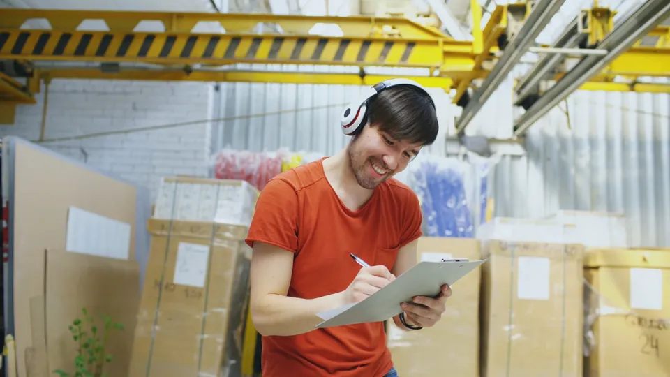 Smiling warehouse worker wearing headphones and writing on clipboard